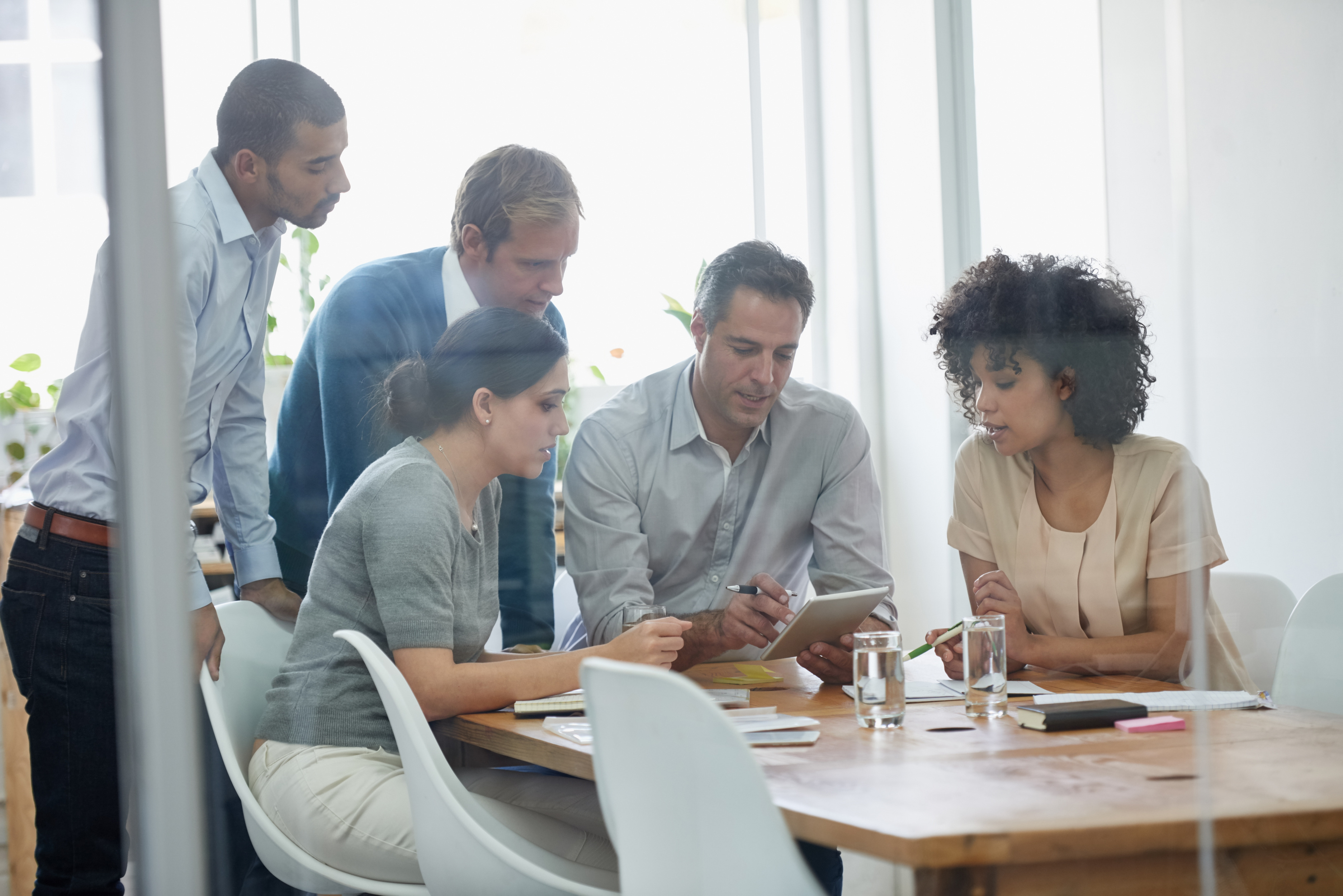 Five colleagues gathered around a bright office table reviewing work on a tablet